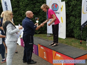 Komendant Miejski Policji we Wrocławiu mł. insp. Rafał Siczek wręczający puchar stojącemu na podium jednej z uczestniczek czwartej edycji Mistrzostw Senioralnych o Puchar Komendanta Miejskiego Policji.