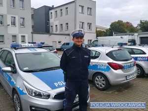 Stojący na tle radiowozów umundurowany policjant ruchu drogowego.