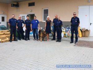 Stojący na wewnętrznym placu wrocławskiego schroniska dla zwierząt policjanci oraz pracownicy schroniska we Wrocławiu, w tle przywieziona przez policjantów karma oraz inne przedmioty dla podopiecznych schroniska.