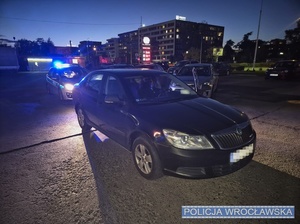 Stojące na jednym z wrocławskich parkingów osobowa skoda oraz oznakowany radiowóz policji włączonymi światłami błyskowymi.