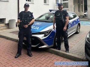 Dwóch umundurowanych policjantów stojących prze budynkiem Komisariatu Policji Wrocław-Fabryczna przed oznakowanym radiowozem Policji.