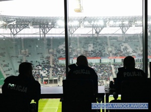 Siedzący w pomieszczeniu stadionu, z widokiem na płytę dowodzący zabezpieczaniem meczu trzej umundurowani policjanci