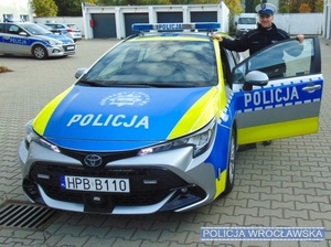 Umundurowany policjant ruchu drogowego stojący na parkingu przy oznakowanym radiowozie.