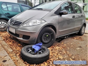 Zdjęcie auta stojącego na parkingu i leżącego obok pełnowymiarowego koła wraz z narzędziami do jego wymiany