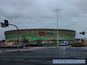 Stojący przy jednym z wrocławskich skrzyżowań na tle Stadionu Miejskiego oznakowany radiowóz Policji z włączonymi sygnałami świetlnymi.