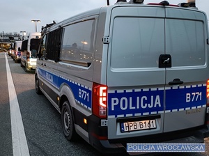 Stojące na drodze wewnętrznej wrocławskiego Stadionu Miejskiego, zaparkowane wzdłuż chodnika, oznakowane radiowozy Policji.