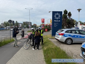Na zdjęciu widać kobietę która stoi obok roweru i jest skontrolowana przez dwóch funkcjonariuszy policji. W tle radiowóz policyjny