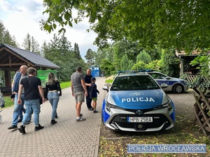 na zdjeciu radiowóz i obok niego policjant z dzieckiem i innymi osobami
