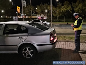 na zdjęciu widać szary samochód i stojącego z tyłu za nim policjanta