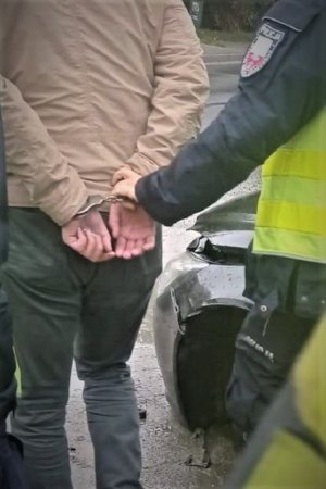 Na zdjęciu kierujący Porsche z kajdankami założonymi na ręce trzymane z tyłu,  prowadzony przez umundurowanego policjanta