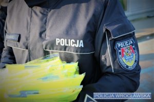 policjant trzyma odblaskowe kamizelki, na rękawie munduru widoczny emblemat Komendy Miejskiej Policji we Wrocławiu