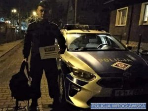 Policjant w mundurze stojący obok oznakowanego radiowozu