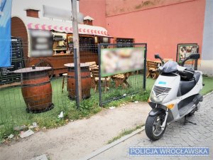 Zdjęcie przedstawia skuter koloru srebrnego. Na drugim planie widoczny jest punkt gastronomiczny koloru brązowego